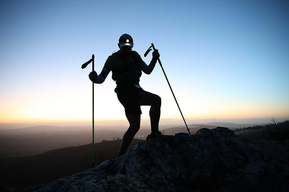 A silhouetted hiker with trekking poles stands atop a rocky ridge at sunrise, outlined against a blue-to-orange sky.